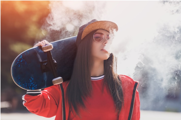 Girl with hat and glasses vaping freebase e-liquid with a skateboard resting on her shoulder